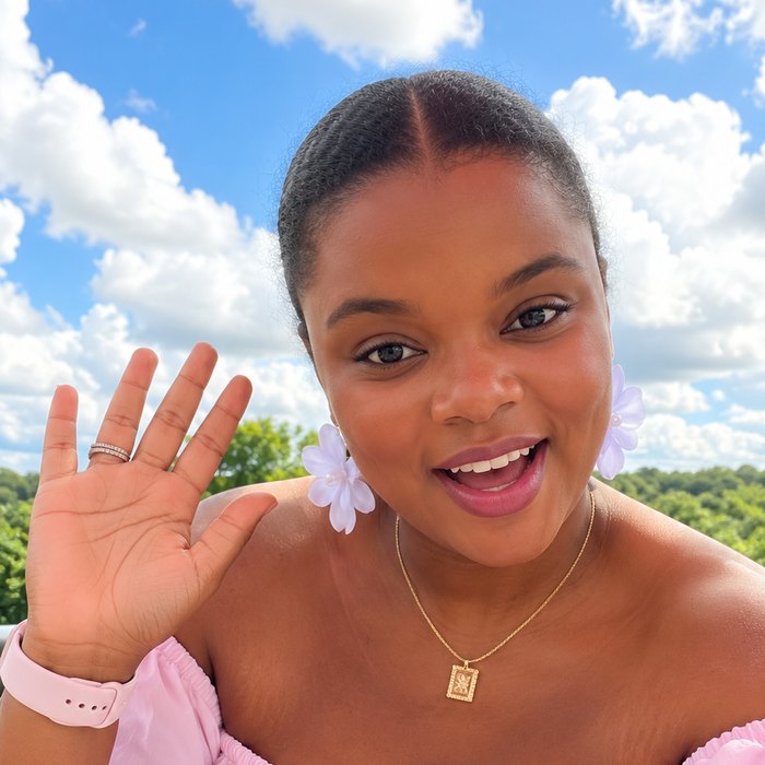Mrs. Roo, the host of My A to Z Trips: a smiling woman in a pink off-the-shoulder top waving hello against a blue sky.
